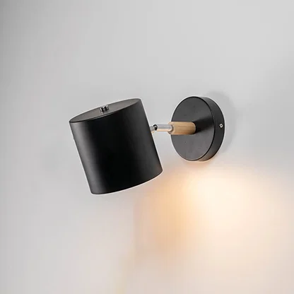 A black wall lamp with light on against a white background showing its cylindrical shade wooden connector and round base creating a clean modern illuminated look.