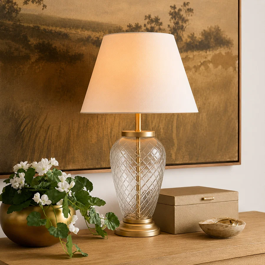Decorative glass table lamp with a white fabric lampshade and gold accents on a wooden console table, styled with a flowering plant and home décor accessories.