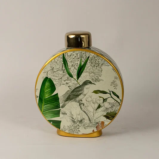 Round ceramic vase with green botanical leaf artwork and gold rim placed on white surface against neutral background showcasing elegant nature inspired home decor styling.