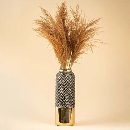 Tall ceramic vase with textured design and golden base displayed with dried pampas stems creating a warm decorative accent against a soft beige backdrop.