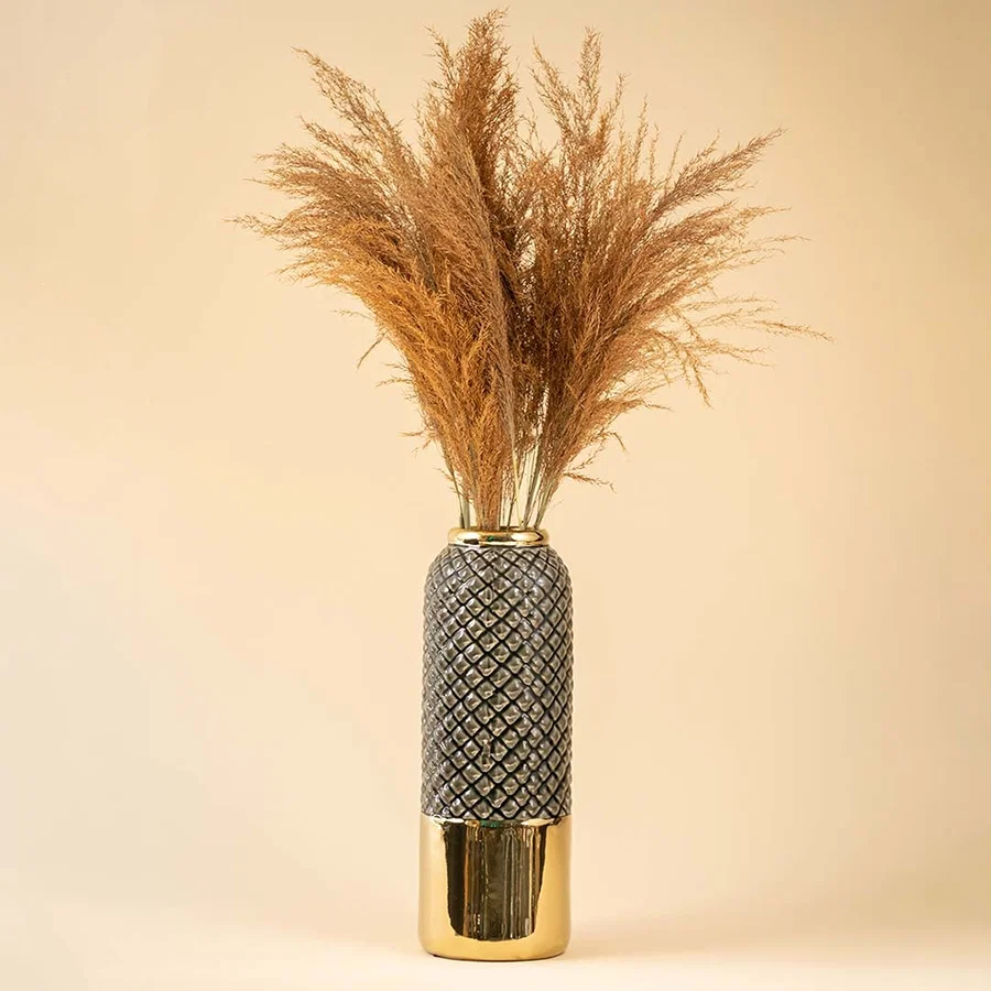 Tall ceramic vase with textured design and golden base displayed with dried pampas stems creating a warm decorative accent against a soft beige backdrop.