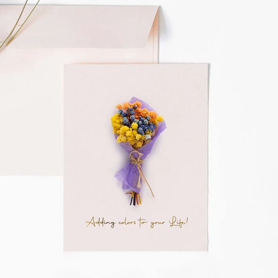A handmade pink greeting card featuring a mini purple and yellow dried flower bouquet next to a matching light pink envelope.