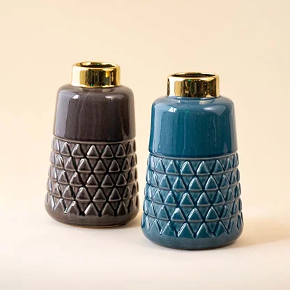 A pair of ceramic vases with geometric patterns and gold rims are shown together one is teal blue and the other is dark grey brown.