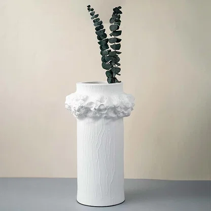 Tall white ceramic vase with textured surface and floral crown detail styled with green leaves for home decor.