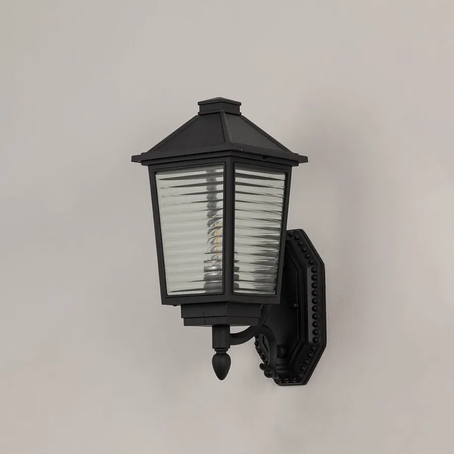 Traditional black metal wall lantern with textured glass panels and ornate bracket switched off on minimal white backdrop.