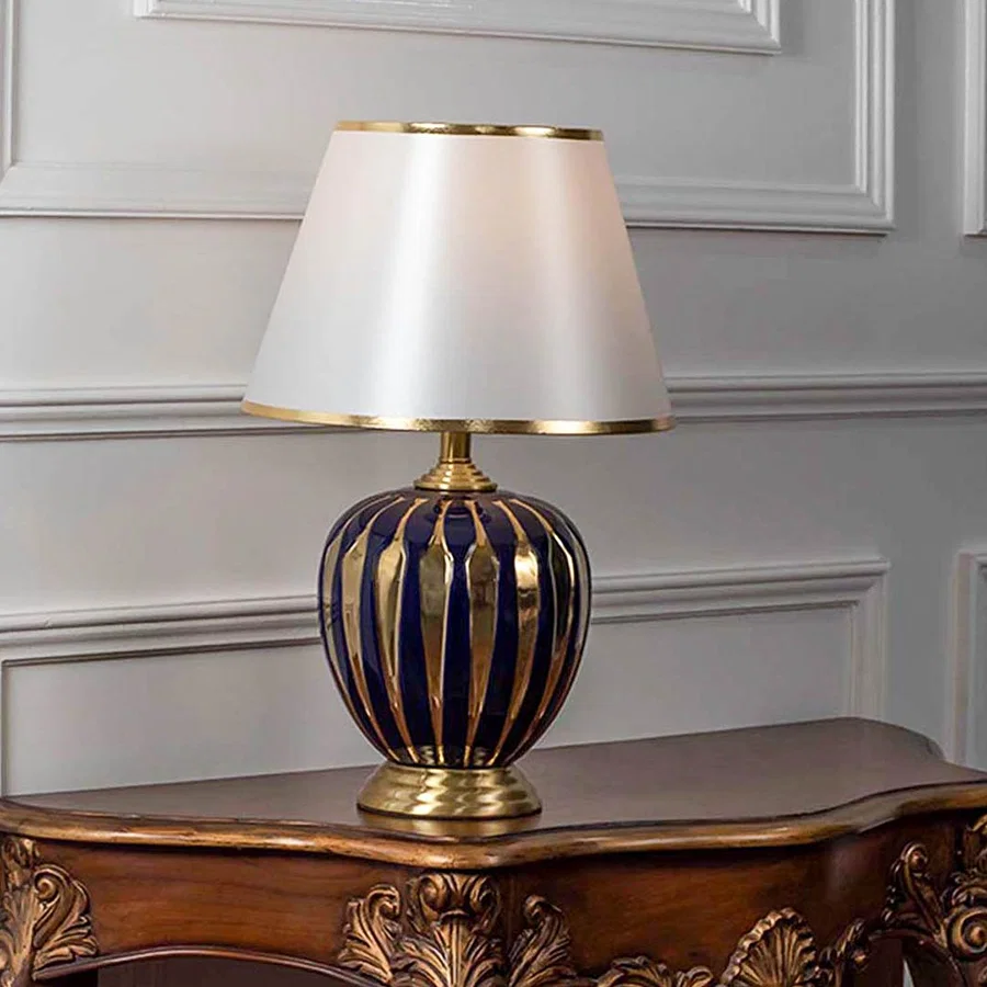 Table lamp with blue glass base and gold accents with white fabric shade on wooden table luxury lighting for living room decor.