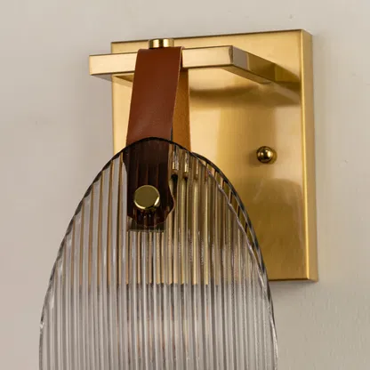 Closeup of gold metal wall light base with ribbed glass shade and leather strap accent on white background for premium interior decor.