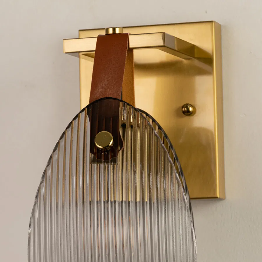 Closeup of gold metal wall light base with ribbed glass shade and leather strap accent on white background for premium interior decor.