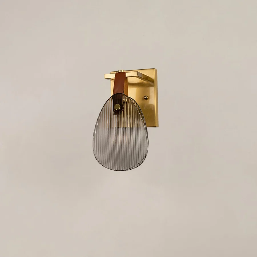 Gold metal wall sconce with ribbed glass and leather strap illuminated on white background for elegant lighting.