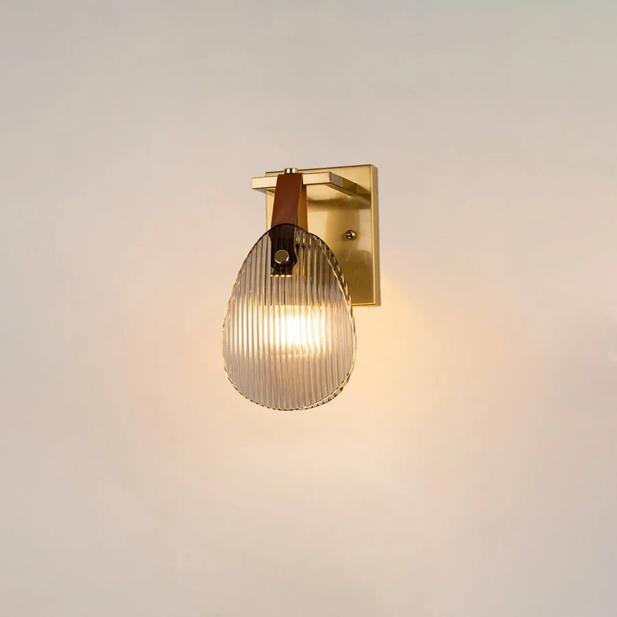 Gold wall light with ribbed glass shade and leather strap glowing on a white background for modern home lighting.