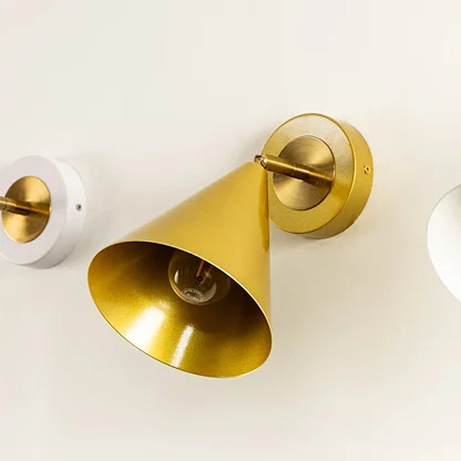 Close-up of a gold cone shaped wall sconce with a visible bulb mounted on a round brass base against a light colored wall.