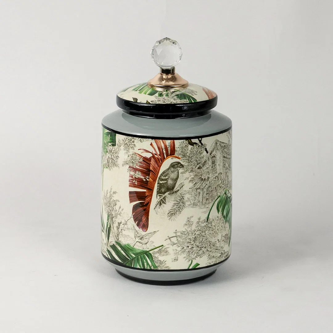 Ceramic jar with tropical leaf and bird artwork and green lid placed on white surface against neutral background highlighting artistic decorative storage styling.