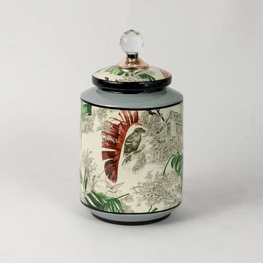 Ceramic jar with tropical leaf and bird artwork and green lid placed on white surface against neutral background highlighting artistic decorative storage styling.