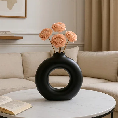 A modern black circular vase styled with soft peach flowers placed on a round white marble table in a warm living room setting creating a calm decorative look.