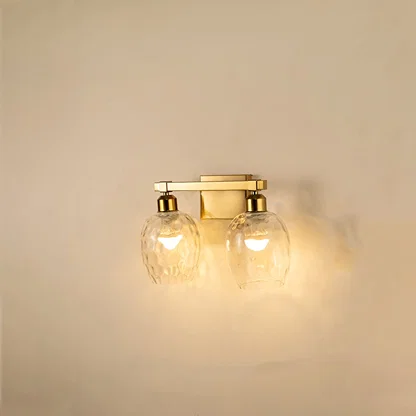 Brushed gold double wall light with hammered clear glass shades glowing on white background adding warm elegant lighting to modern interiors.
