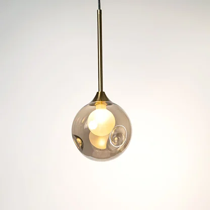 Clear glass globe pendant light with gold holder and visible bulb hanging against neutral wall background highlighting elegant contemporary lighting design.