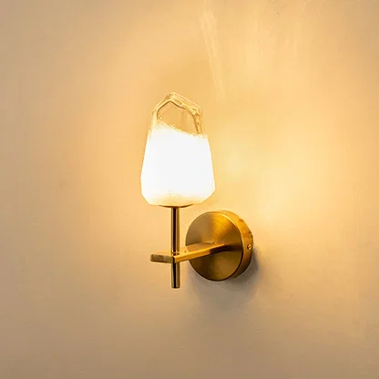 Brushed gold wall light with crystal cut frosted glass shade glowing on white background adding soft warm lighting to modern interior spaces.