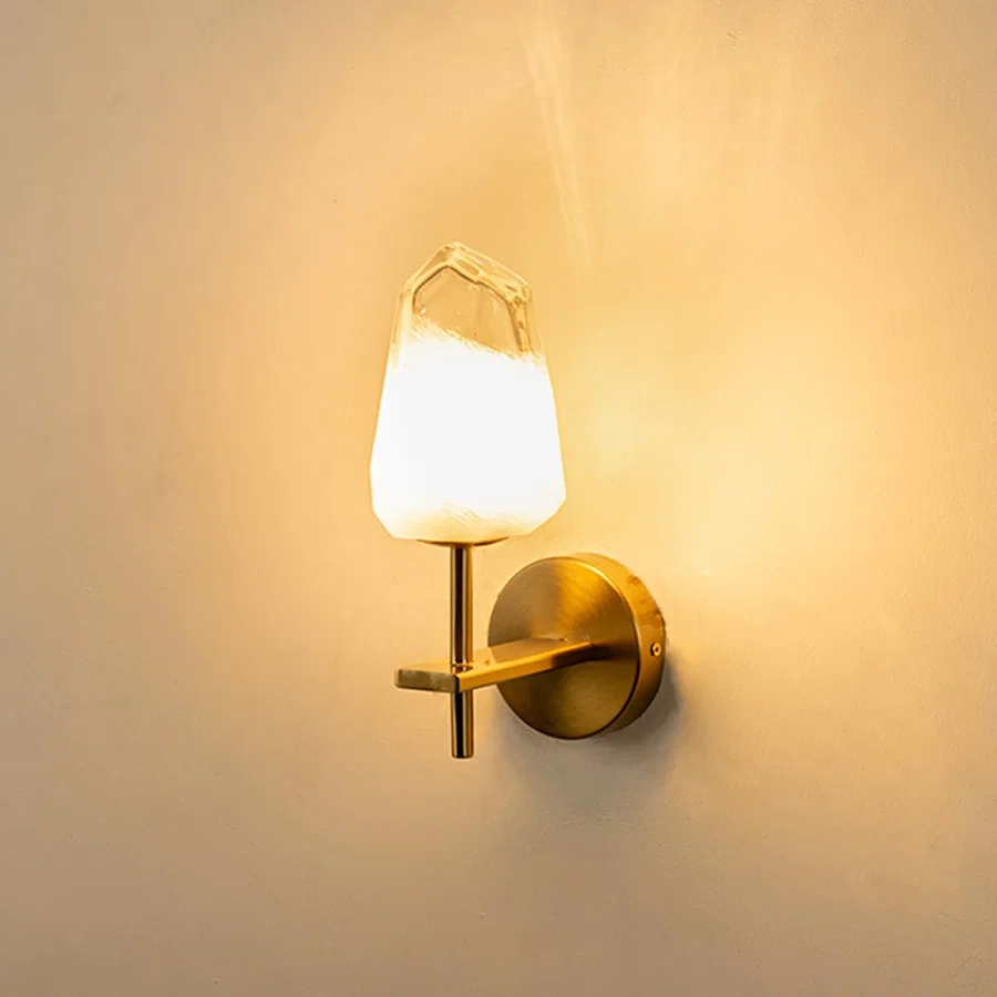 Brushed gold wall light with crystal cut frosted glass shade glowing on white background adding soft warm lighting to modern interior spaces.