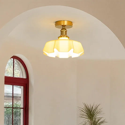 A semi-flush ceiling light fixture with a warm yellow glass shade and a gold-toned brass ceiling mount, shown installed in an arched interior space with a red-framed window and a plant.