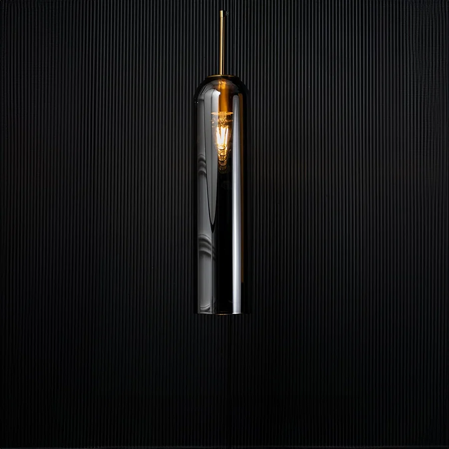 A modern silver cylindrical pendant light against a deep black background with the light turned on casting a warm glow and highlighting its reflective finish.