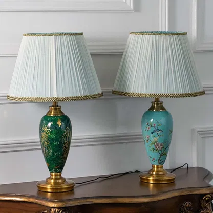Emerald green ceramic table lamp featuring intricate gold leaf detailing and a pleated white shade turned off on a white background highlighting its glossy artistic finish.