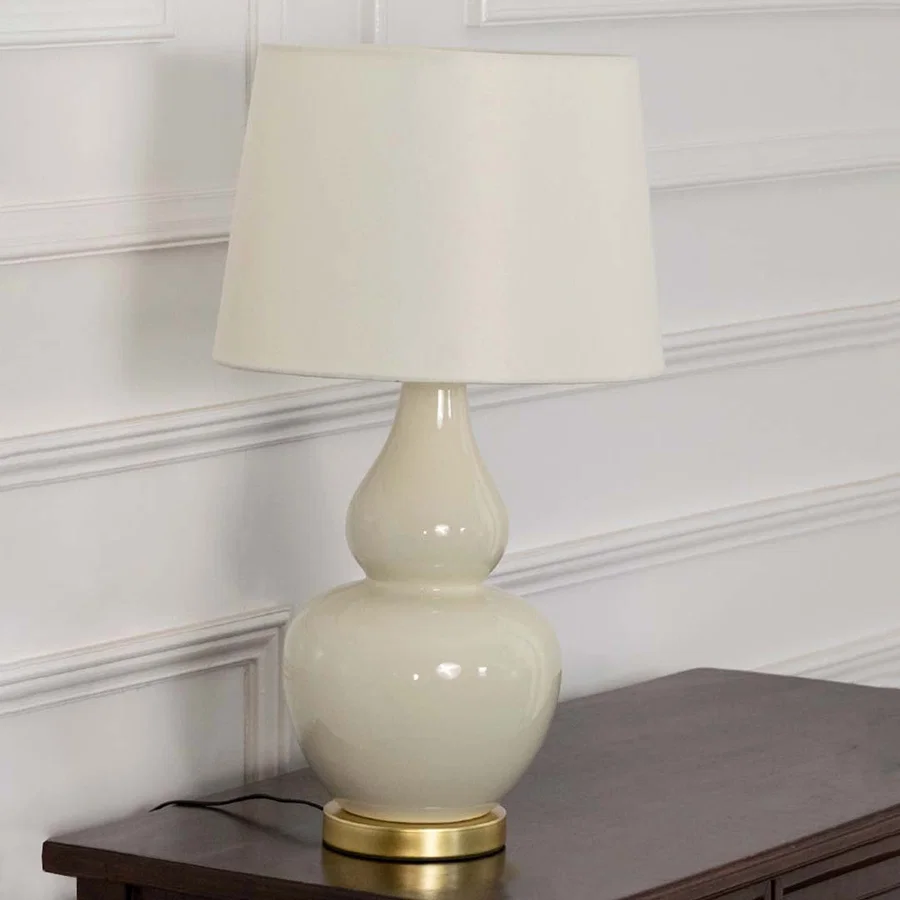 White shade beige ceramic table lamp on wooden side table with light off and white panel wall for clean modern décor.