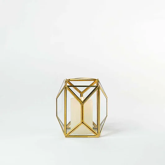 Gold metal and glass geometric lantern shown on a white background with candle on highlighting its modern structure and warm inviting glow.