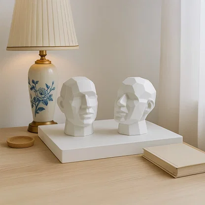 Two white geometric face sculptures displayed on a wooden table beside a lamp adding modern artistic touch to decor.