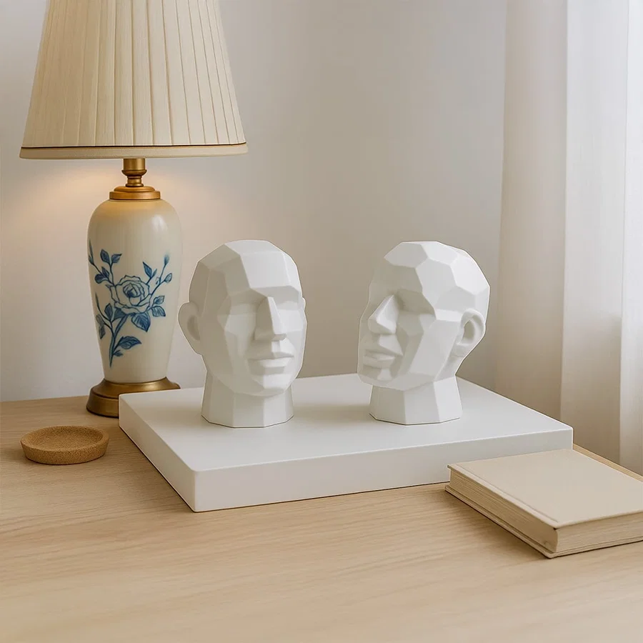 Two white geometric face sculptures displayed on a wooden table beside a lamp adding modern artistic touch to decor.