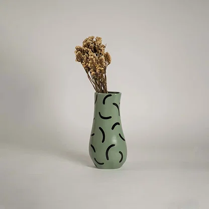 Tall green ceramic vase with black curved patterns holding dried flowers placed on a wooden chest to enhance a cozy vintage themed room.
