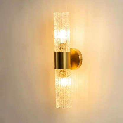 A gold wall sconce with two textured cylindrical glass shades shown with lights on against a white background highlighting its warm ambient glow and vertical design.