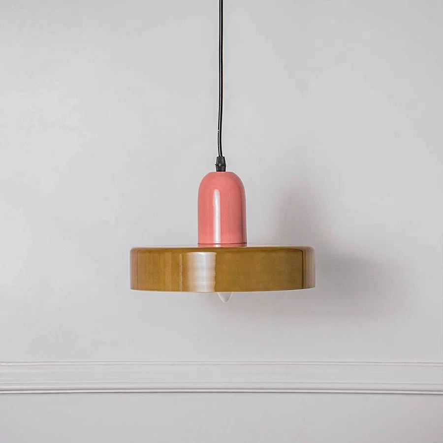 Decorative pendant light with pink holder and wooden disc shade placed against neutral wall background showcasing playful contemporary lighting design.