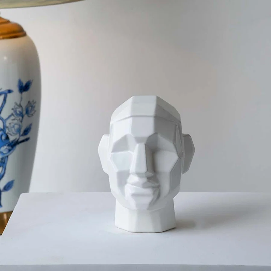 Modern geometric face sculpture displayed on a minimal white platform with soft natural light creating subtle shadows and depth.