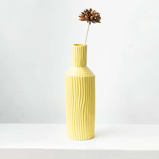 A single brown dried decorative flower pod stem arranged inside a textured pastel yellow ceramic vase on white backdrop.