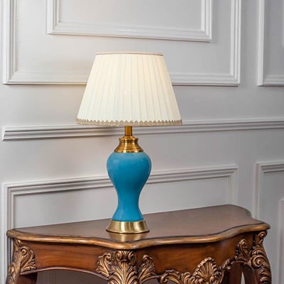 Blue ceramic table lamp turned on with pleated fabric shade and brass detailing displayed on white background.