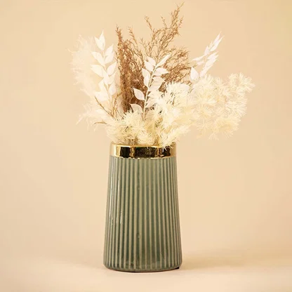 Elegant ribbed green ceramic vase with golden rim filled with soft white and beige dried flowers placed against a warm beige background for a sophisticated home decor look.