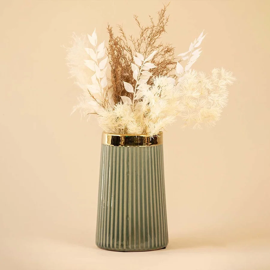 Elegant ribbed green ceramic vase with golden rim filled with soft white and beige dried flowers placed against a warm beige background for a sophisticated home decor look.