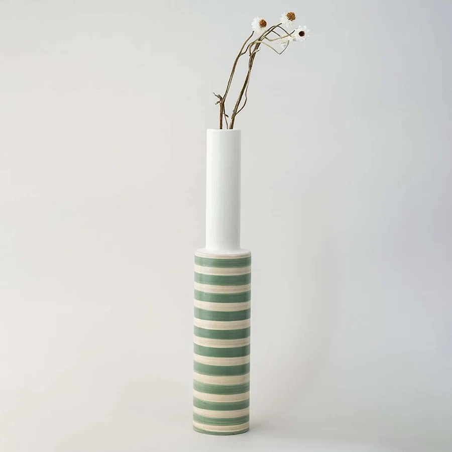 A long neck striped vase captured in bright natural light on a simple white platform showcasing its sleek artistic shape and soft green tones in a modern lifestyle scene.