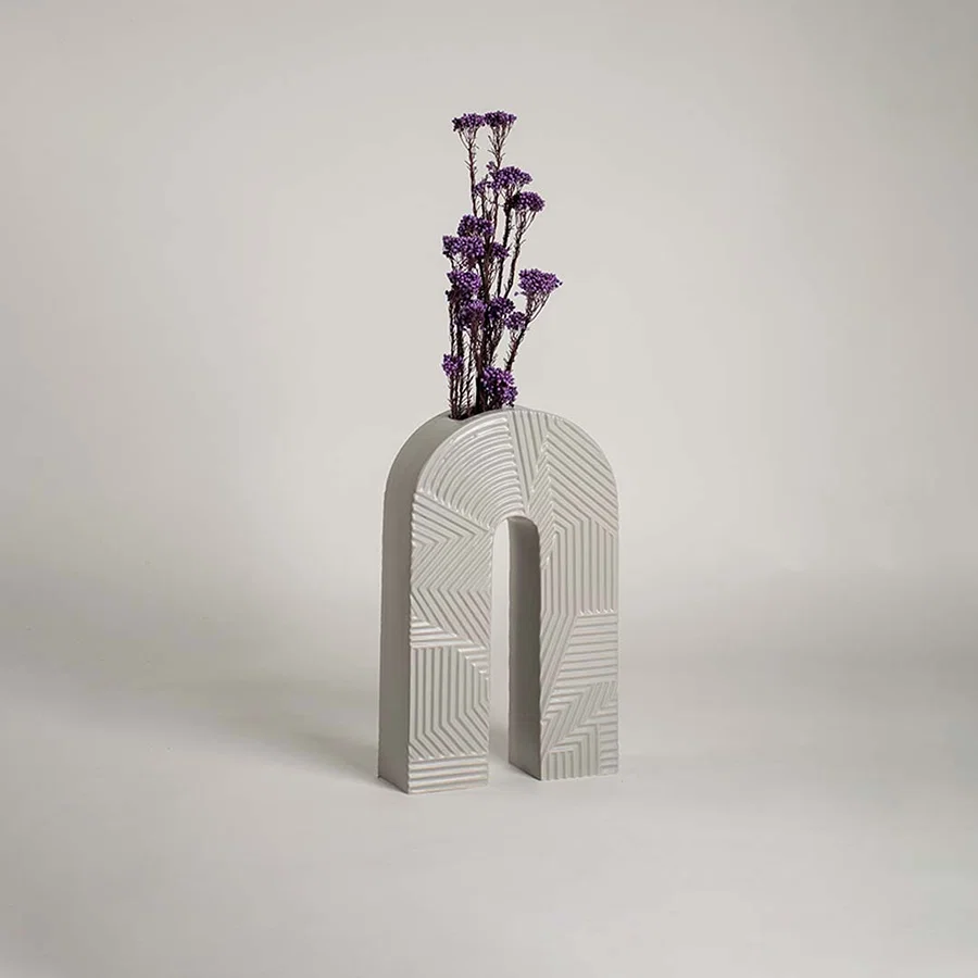 White ceramic arch vase with geometric texture holding purple flowers on a wood table next to a boucle chair for modern decor.