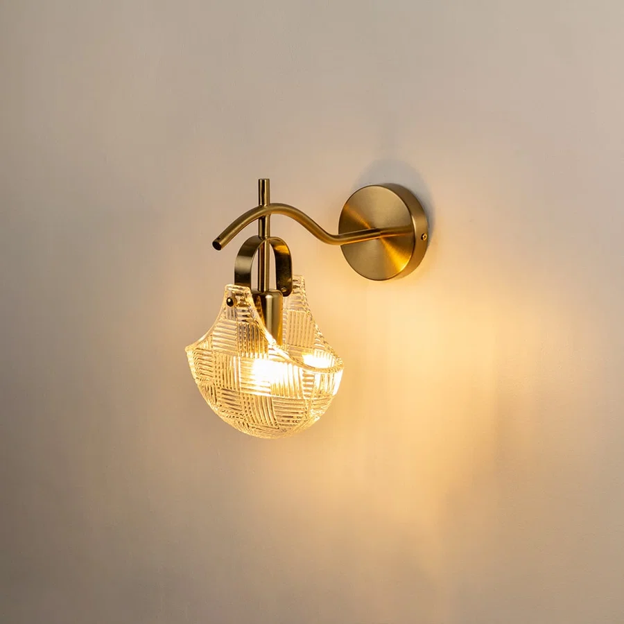 A gold metal wall light with a patterned clear glass shade shown with lights on against a white background highlighting its warm soft glow and elegant shape.