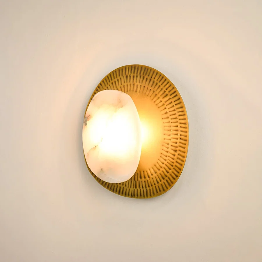 Contemporary alabaster wall sconce illuminated on white background with textured brass plate offering a soft  warm glow and sophisticated appeal.