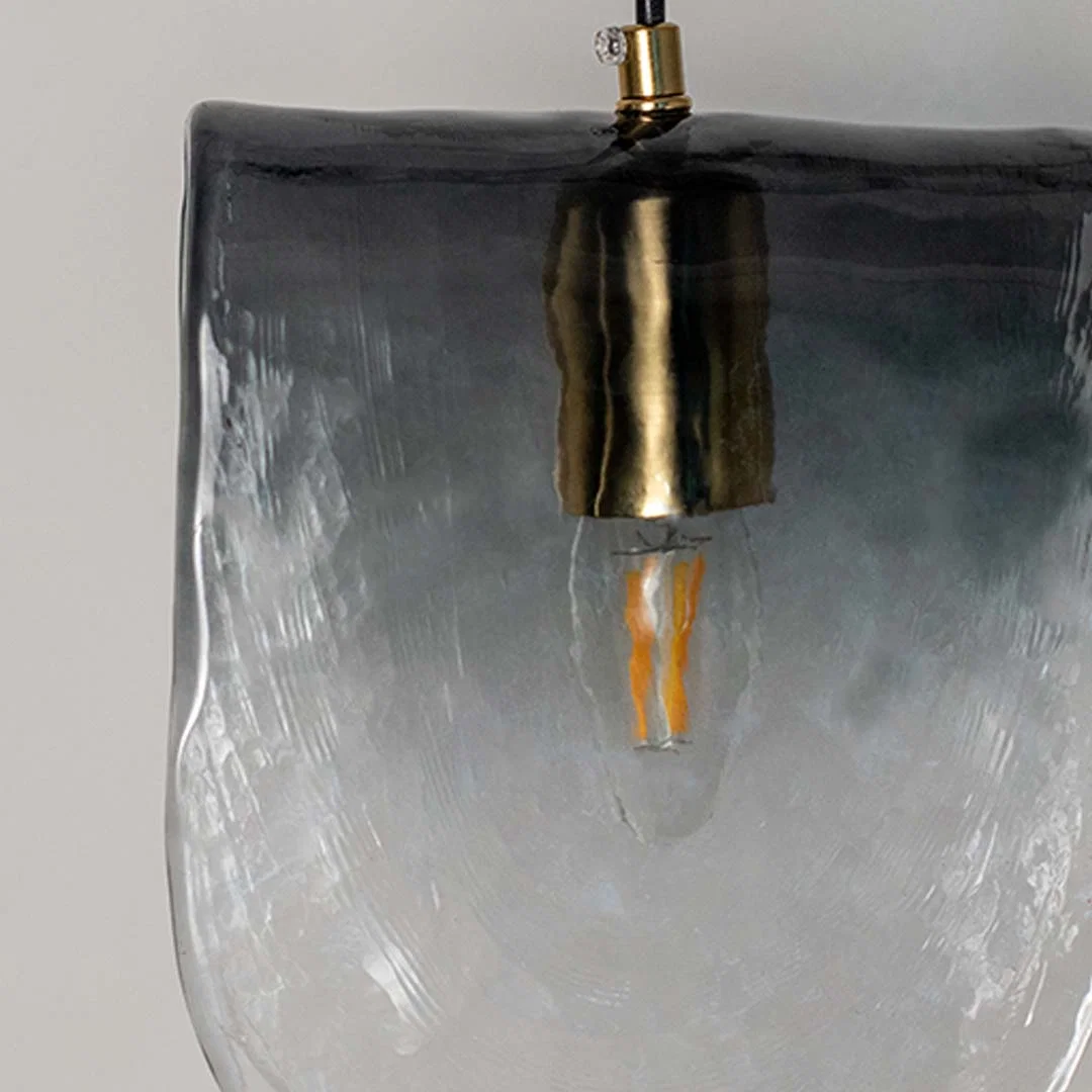Frosted glass pendant light with smoky gradient finish and gold fitting placed against neutral wall background closeup highlighting modern artistic lighting decor styling.