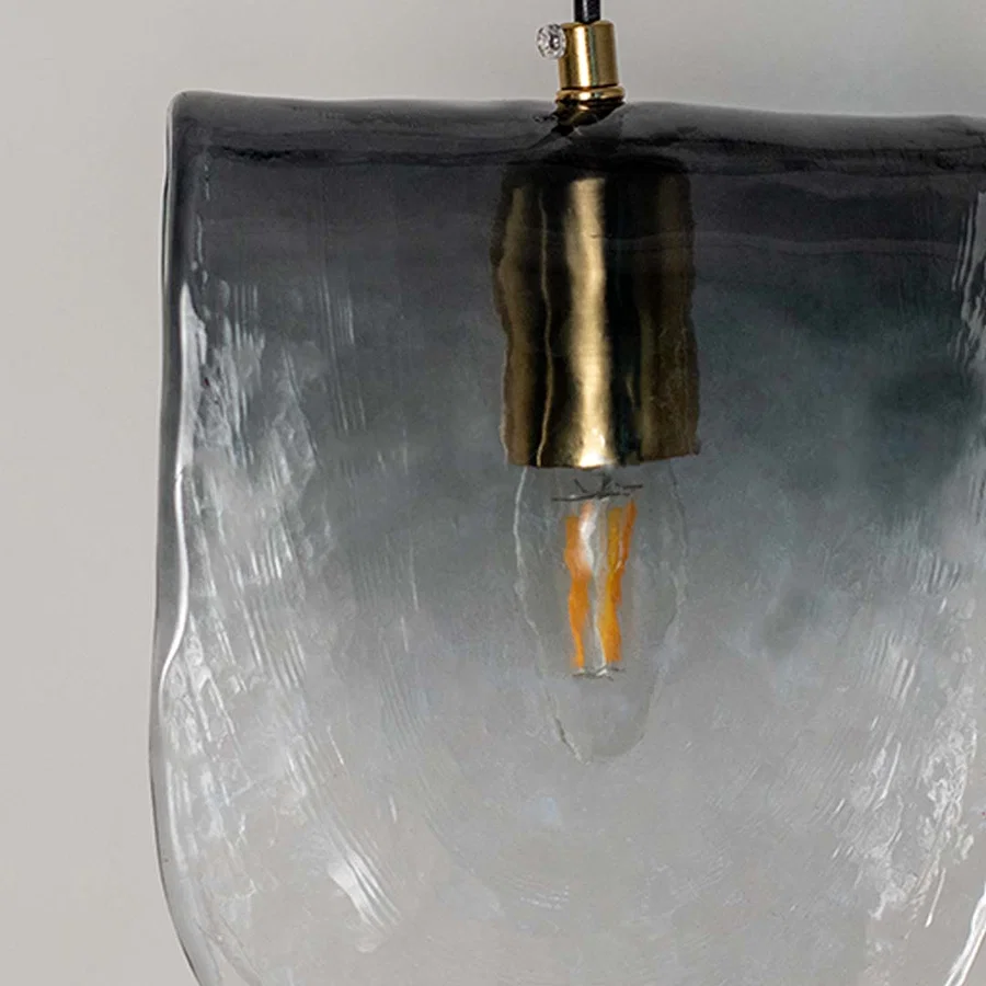 Frosted glass pendant light with smoky gradient finish and gold fitting placed against neutral wall background closeup highlighting modern artistic lighting decor styling.