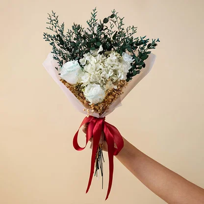 Hand holding an elegant white preserved hydrangea and green eucalyptus leaves bouquet tied with a red ribbon backdrop.