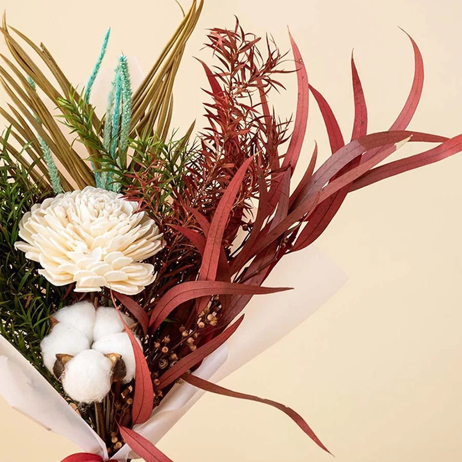 Closeup of dried bouquet with shola flower cotton stem palm leaf and red eucalyptus wrapped in white paper with satin ribbon.