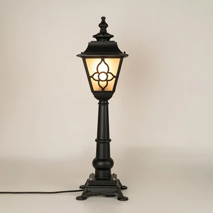 A traditional outdoor pillar lamp displayed on a white background with the light on showing its frosted glass panels ornate metal pattern and tall sculpted post design.