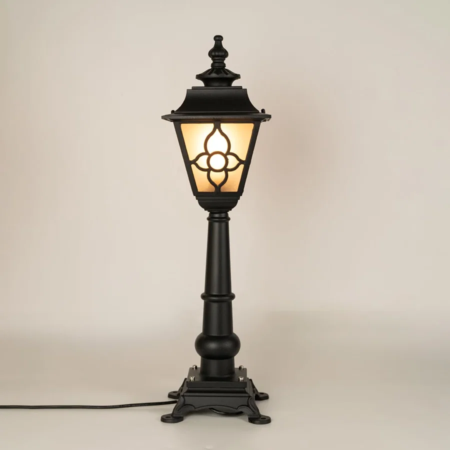 A traditional outdoor pillar lamp displayed on a white background with the light on showing its frosted glass panels ornate metal pattern and tall sculpted post design.
