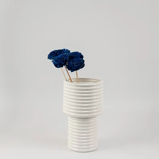 Royal blue handmade sola wood flower stems arranged inside a modern white ribbed ceramic vase for bedroom tabletop decoration.