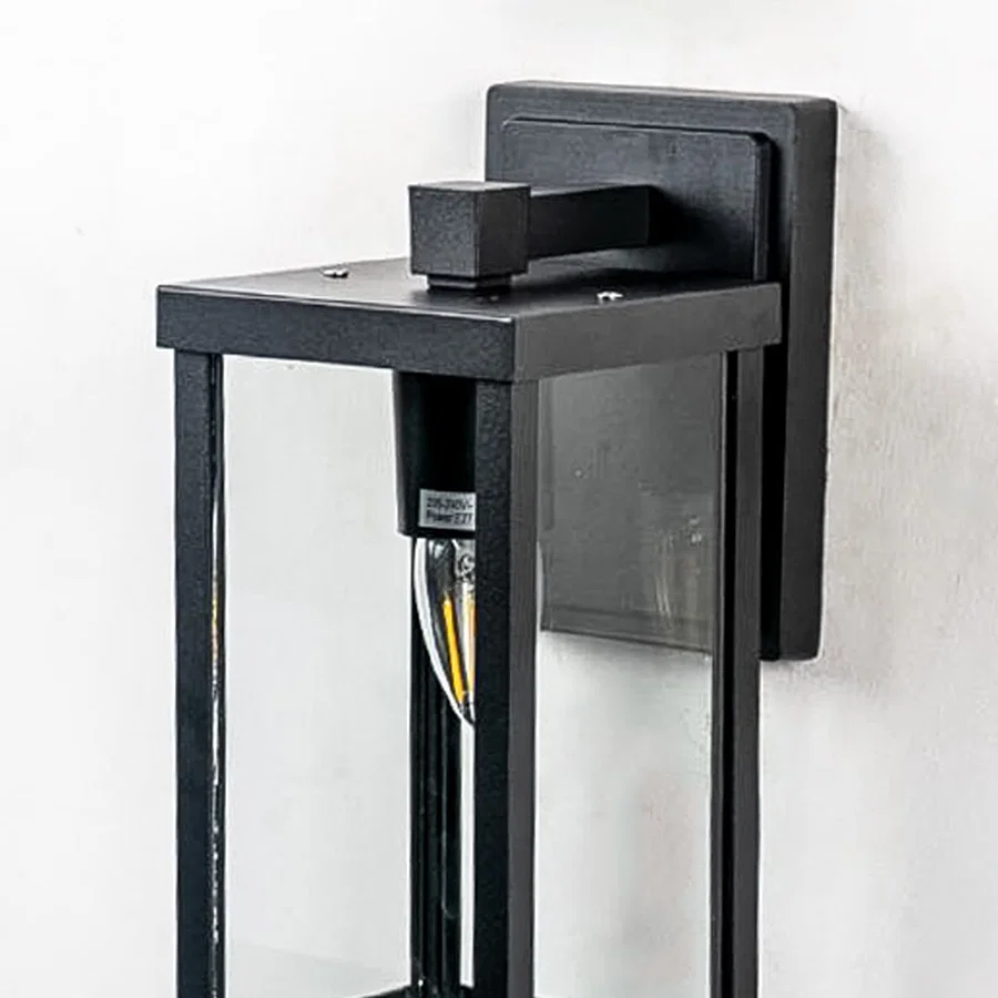Closeup of outdoor wall light with black frame and transparent glass shade highlighting warm glowing bulb design.
