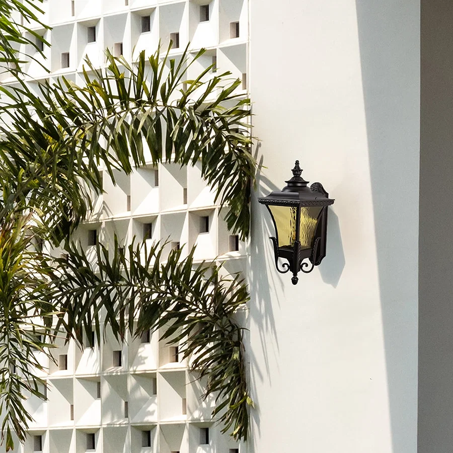 Bronze metal outdoor wall light lifestyle image with amber glass shade installed on a white villa facade for decor.